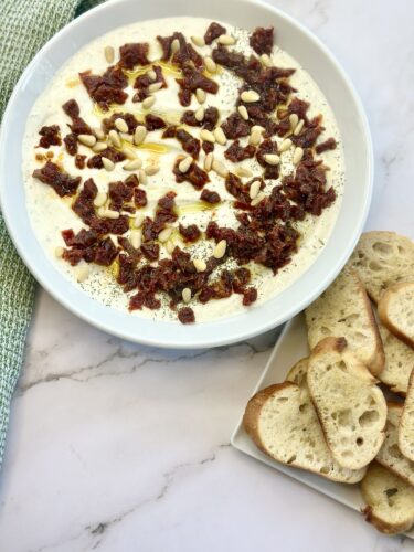 A bowl of creamy dip topped with sun-dried tomatoes, pine nuts, and herbs, accompanied by slices of toasted bread on a plate.