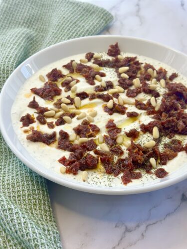 A bowl of yogurt topped with pine nuts and minced dried tomatoes, garnished with olive oil and herbs, placed on a marble surface next to a green textured napkin.