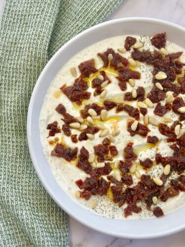 A bowl of creamy yogurt topped with chopped sun-dried tomatoes, drizzled with olive oil, and sprinkled with pine nuts and herbs, set on a marble surface next to a textured green napkin.