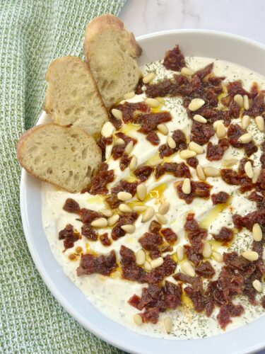 A white bowl filled with a creamy spread topped with sun-dried tomatoes, pine nuts, and drizzled with olive oil, accompanied by slices of crusty bread, resting on a green cloth.