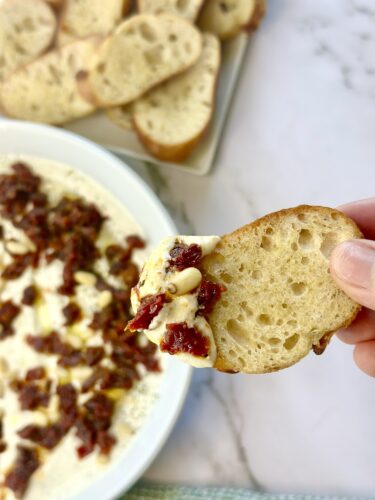 A hand holding a slice of toasted bread topped with a creamy dip and sun-dried tomatoes, with a bowl of dip and additional bread slices in the background.