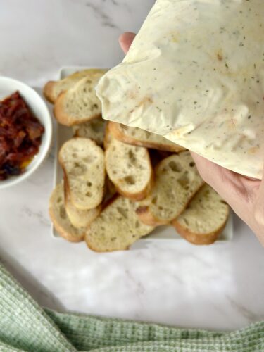 A hand holding a package of creamy dip over a platter of sliced bread, with a small bowl of chopped bacon in the background.