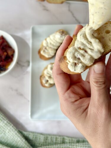 A hand holding a piece of bread topped with creamy spread, with additional pieces of bread and a small bowl of toppings in the background.
