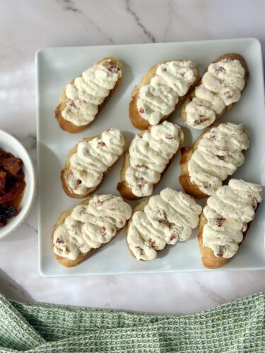 A platter of cream-topped crostini arranged on a white plate, with a small bowl of sun-dried tomatoes on the side.