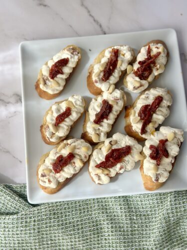 A platter of toasted crostini topped with creamy spread and sun-dried tomatoes, arranged neatly on a light-colored plate.