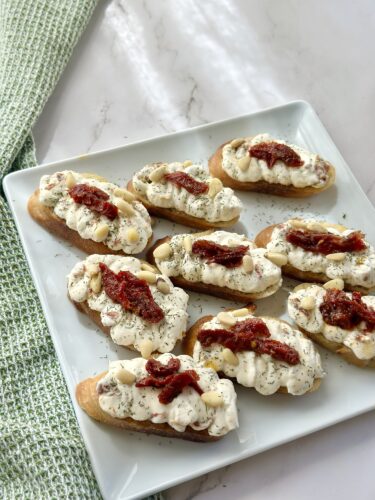A platter of toasted bread slices topped with cream cheese, herbs, and sun-dried tomatoes, arranged neatly on a light-colored plate.