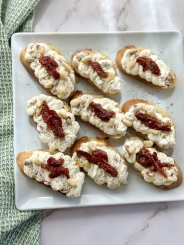 A platter of crostini topped with creamy cheese, sun-dried tomatoes, and almonds, arranged neatly on a white square plate.