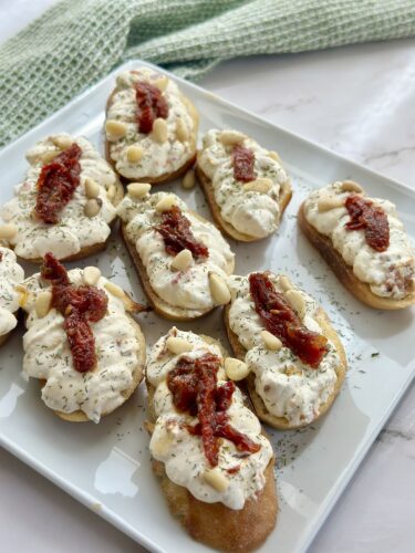 A close-up of a plate filled with toasted bread slices topped with creamy spread, sun-dried tomatoes, pine nuts, and herbs.