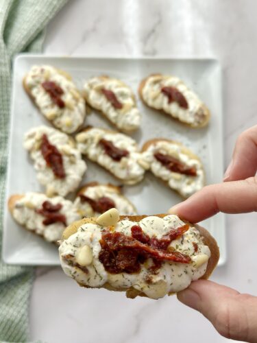 A hand holding a slice of bread topped with creamy spread, sun-dried tomatoes, and herbs, with a plate of similar appetizers in the background.